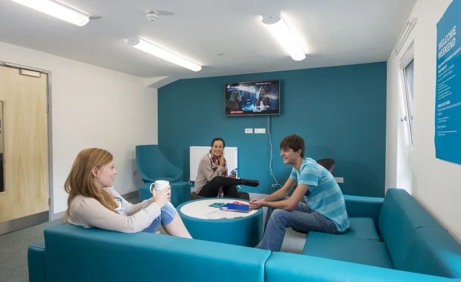 A group of students sit and talk whilst watching TV in a shared common room.