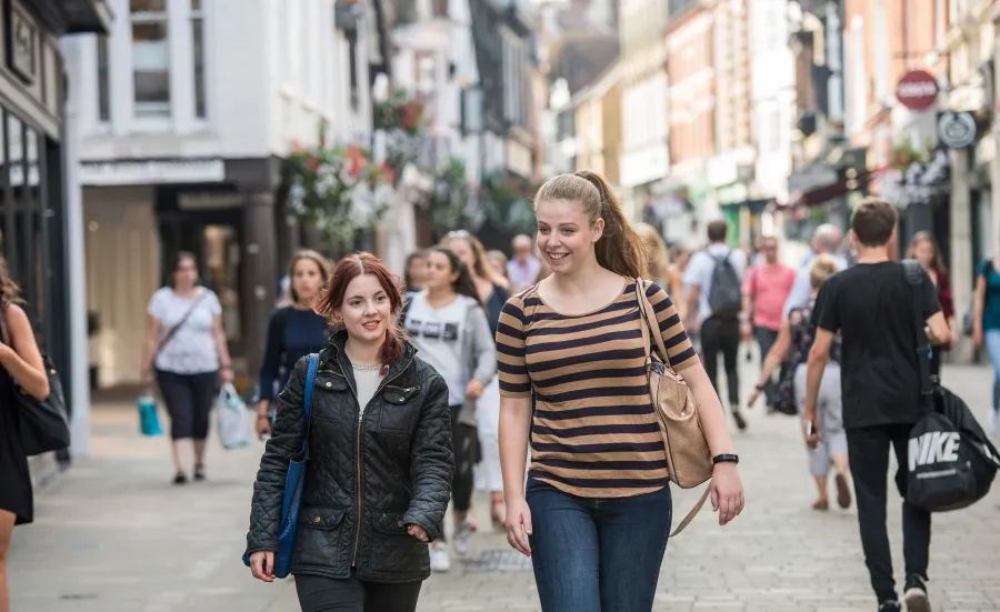 Two students talking whilst walking down a busy, cobbled shopping street.