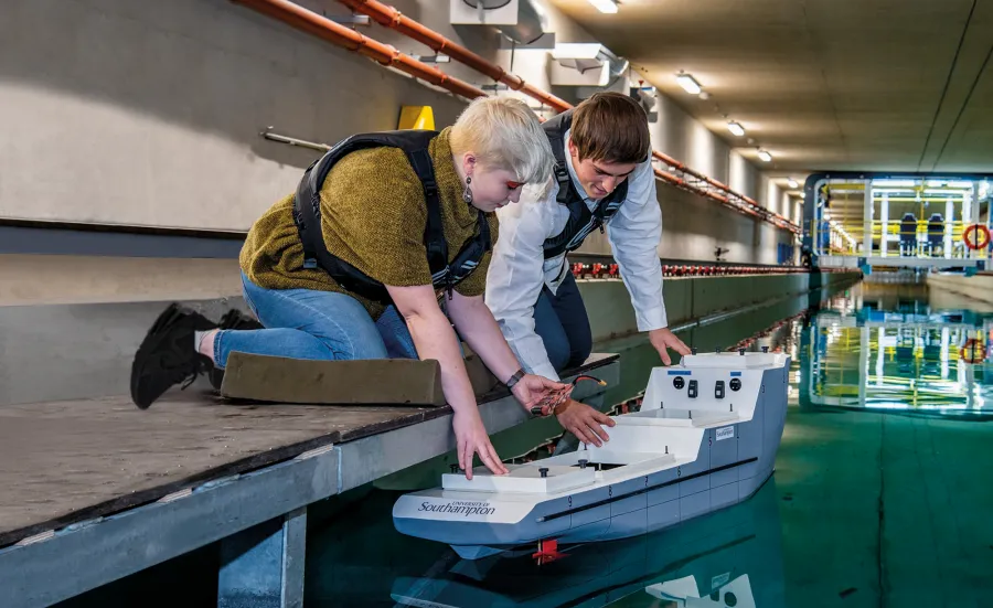 A student crouches down to place a model boat into the water of the university towing tank, a researcher at her side