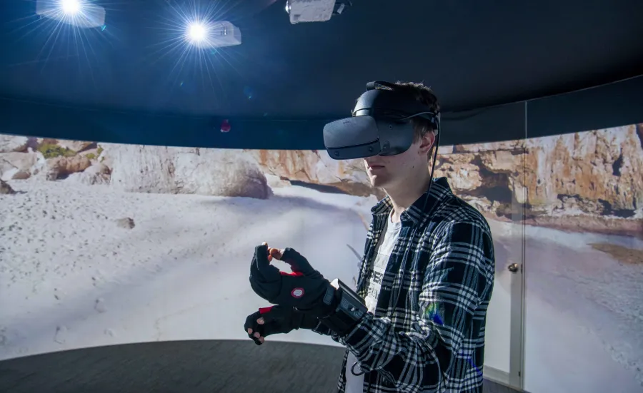 A student wearing a virtual reality headset and gloves, standing in front of a 360 degree video wall displaying what he can see.
