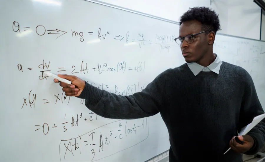 A maths teacher writes on a white board and looks intently at the equations. We imagine he is in front of a classroom of pupils.