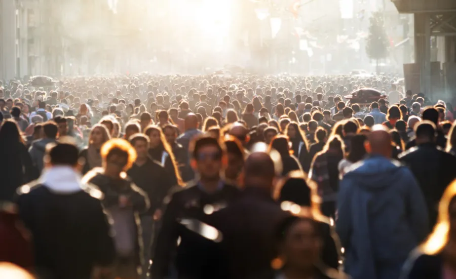 A large crowd of people spans from the foreground to the background. The people closer to us are picked out as indviduals because of a low, bright sunlight.