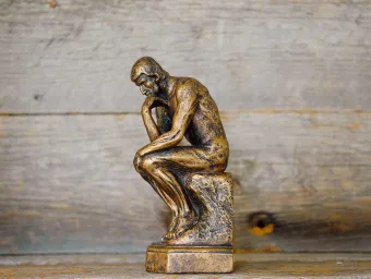 A small replica of 'The Thinker' - a bronze sculpture by Auguste Rodin depicting a man hunched over on a rock in contemplation - set against a backdrop of wood panels.