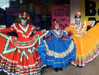 Three dancers wearing brightly coloured Mexican dresses and headgear pose for a photograph