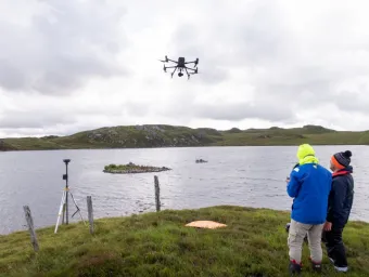 Two archaeologists guide a drone into the air over a grey stretch of water between grassy, low-lying islands. They are well wrapped up against the cold. 